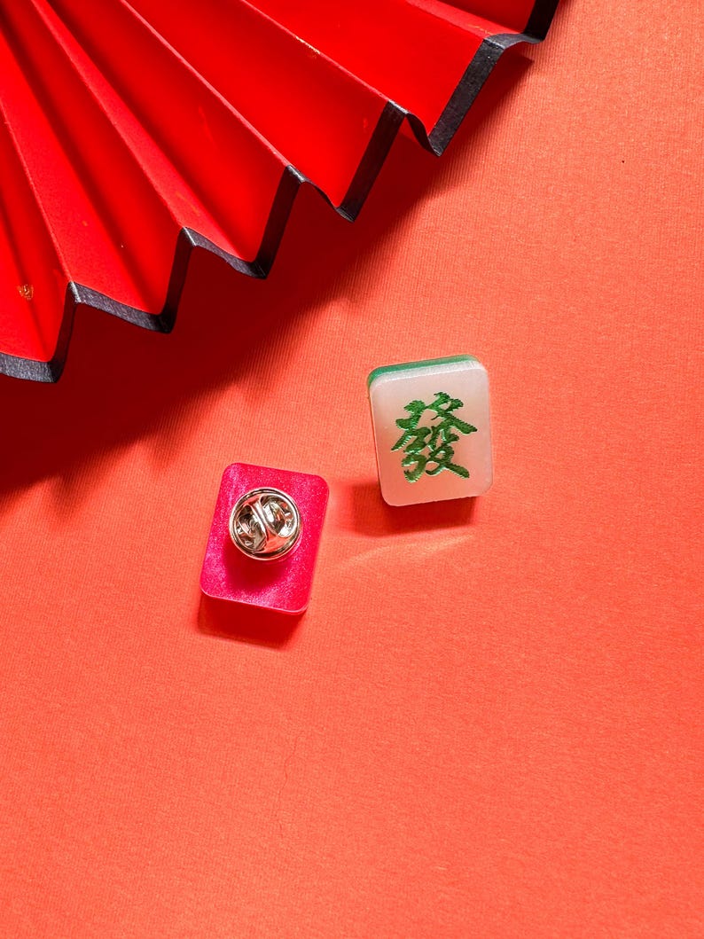 May include: Two small square pins with a red and white background. One pin features a green Chinese character, the other pin has a silver pin back.