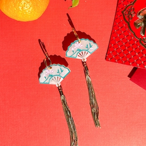 May include: A pair of fan-shaped earrings with gold hoops and long gold tassels. The earrings feature a white background with a teal border and a pink lotus flower design. The earrings are on a red background with a mandarin orange and a red envelope.
