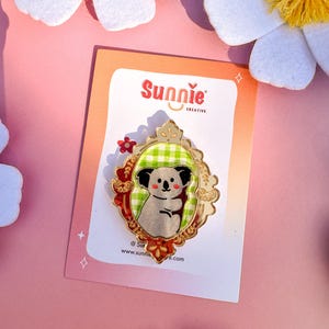 May include: A gold-colored enamel pin featuring a koala bear wearing a green and white checkered shirt, framed in a gold-colored ornate frame with red accents. The pin is on a white card with the text "Sunnie Creative" and a website address.