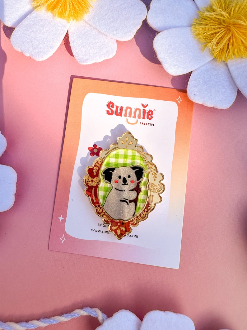 May include: A gold-colored enamel pin featuring a koala bear wearing a green and white checkered shirt, framed in a gold-colored ornate frame with red accents. The pin is on a white card with the text "Sunnie Creative" and a website address.