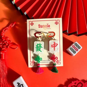 Puede incluir: Un par de pendientes con fichas de mahjong chinas verdes y rojas con detalles dorados. Las fichas están decoradas con caracteres chinos y los pendientes tienen borlas. Los pendientes se muestran en una tarjeta blanca con el nombre de la marca "Sunnie Creative" impreso en ella.