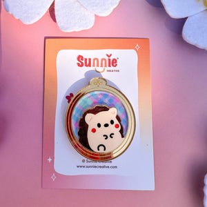 May include: A gold-colored pin with a round frame featuring a cartoon hedgehog with pink cheeks. The frame has a blue and purple background with a glittery effect. The pin is on a pink background with white flowers.