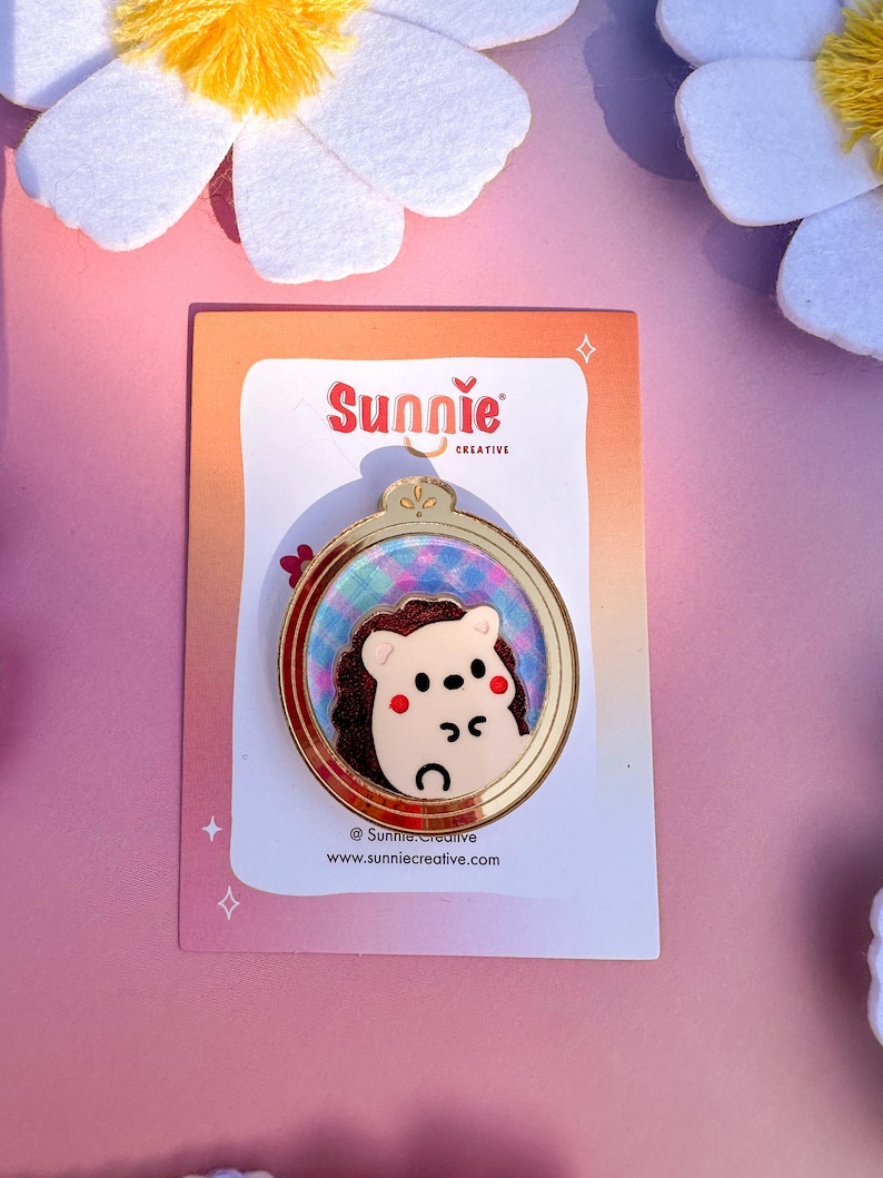 May include: A gold-colored pin with a round frame featuring a cartoon hedgehog with pink cheeks. The frame has a blue and purple background with a glittery effect. The pin is on a pink background with white flowers.