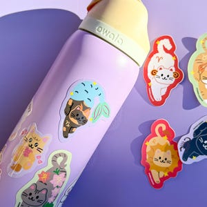 May include: A light purple Owala water bottle adorned with cat-themed stickers. The stickers feature cartoon cats representing zodiac signs. The bottle has a yellow handle and lid. The stickers are in various colors, including orange, gray, and black.