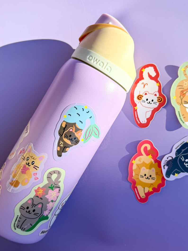 May include: A light purple Owala water bottle adorned with cat-themed stickers. The stickers feature cartoon cats representing zodiac signs. The bottle has a yellow handle and lid. The stickers are in various colors, including orange, gray, and black.