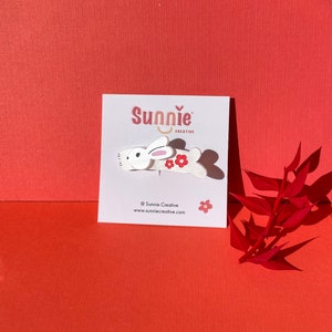 Peut inclure: Barrette pour cheveux en bois blanc en forme de lapin avec des fleurs rouges. La barrette est sur une carte blanche avec le texte "Sunnie Creative" et une adresse de site web.