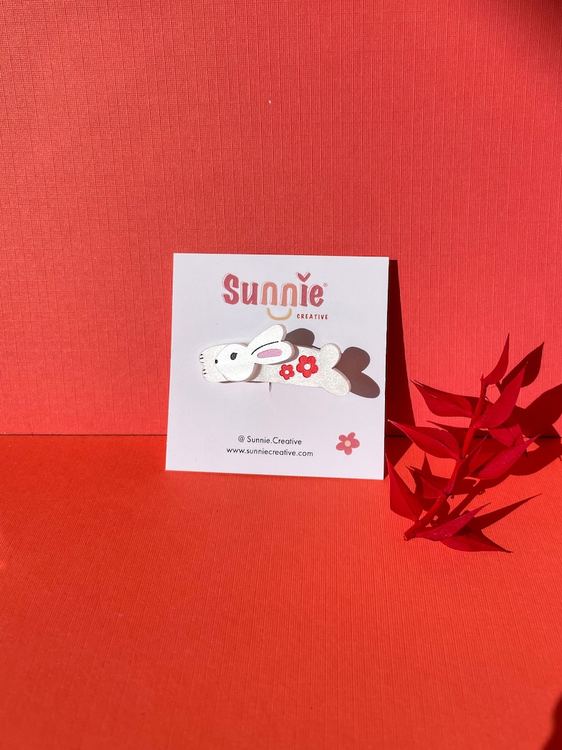 Peut inclure: Barrette pour cheveux en bois blanc en forme de lapin avec des fleurs rouges. La barrette est sur une carte blanche avec le texte "Sunnie Creative" et une adresse de site web.