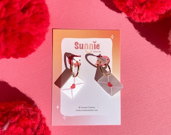 May include: A pair of white and pink envelope earrings with a red heart on each envelope. The earrings are on a white card with the text "Sunnie Creative" and a website address.