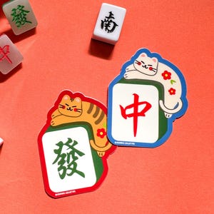 May include: Two colorful stickers featuring a white cat and an orange tabby cat resting on mahjong tiles. The tiles feature Chinese characters, "中" and "發".