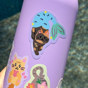 May include: A purple water bottle adorned with several stickers. The primary sticker features a cat with a mermaid tail, in shades of brown, blue, and green. Other stickers include a yellow cat with a pink bow and a gray cat with pink flowers.