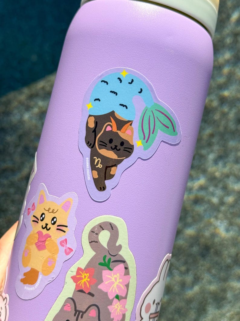 May include: A purple water bottle adorned with several stickers. The primary sticker features a cat with a mermaid tail, in shades of brown, blue, and green. Other stickers include a yellow cat with a pink bow and a gray cat with pink flowers.