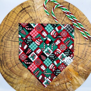 May include: A triangular pet bandana featuring a festive patchwork pattern of red, green, black, and white squares. The design includes holiday icons such as snowflakes, candy canes, Christmas trees, snowmen, stockings, and gift boxes.