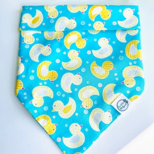 May include: A triangular pet bandana in bright turquoise fabric featuring a repeating pattern of yellow rubber ducks with grey patterned wings and small pink beaks, accented by tiny light pink and blue dots, with a white brand label attached.