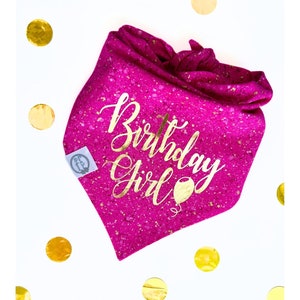 May include: A fuchsia dog bandana with gold flecks and the text "Birthday Girl" in gold script. A gold balloon graphic is below the text. The bandana is on a white surface with gold confetti.