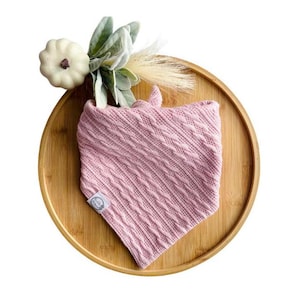Peut inclure: Un bandana en tricot rose pour bébé avec un motif en tricot torsadé. Le bandana est sur une surface en bois.