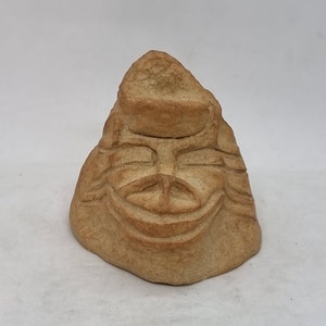 Puede incluir: Una pequeña escultura de cerámica hecha a mano de una cara con una amplia sonrisa. La escultura está hecha de arcilla marrón claro y tiene una superficie rugosa y texturizada.