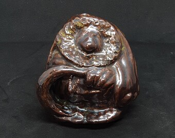 May include: A small ceramic figurine of a brown gnome holding a curved object in its hands. The gnome is wearing a brown hat and a brown robe.
