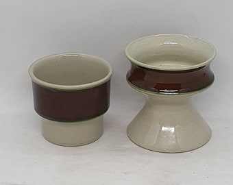 以下が含まれることがあります： 茶色と緑のバンドが付いたセラミック製のカップ2つ。左側のカップは標準的な円筒形。右側のカップは背が高く、砂時計の形をしている。