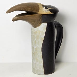 May include: A ceramic pitcher shaped like a toucan with a brown beak, black and white body, and a white eye. The pitcher has a handle and a lid.