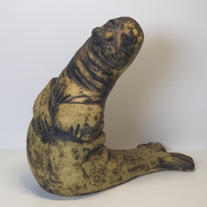 Könnte beinhalten: Eine Keramik-Skulptur eines Seehundes mit brauner und gelber Glasur. Der Seehund sitzt auf seinen Hinterflossen, die Vorderflossen vor der Brust gekreuzt.