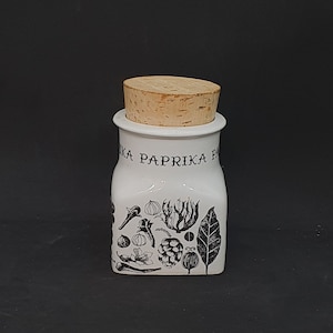 Puede incluir: Tarro de especias de cerámica blanca con tapa de corcho e ilustraciones en blanco y negro de pimientos de paprika y otras especias. El tarro dice "Paprika" en la parte delantera.