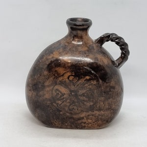 May include: A brown ceramic jug with a twisted handle and a circular design on the front. The jug has a speckled brown glaze.