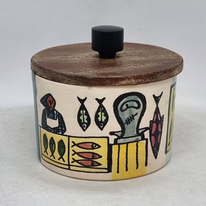 Puede incluir: Un tarro de cerámica con tapa de madera y pomo negro. El tarro está decorado con una ilustración colorida de una escena de mercado de pescado.