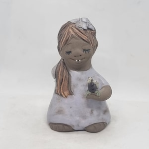 Pode incluir: Uma figurinha de cerâmica de uma menina com cabelo castanho e um vestido branco. Ela está segurando uma pequena flor na mão e tem uma flor branca na cabeça.