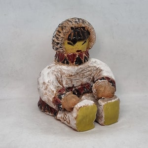 May include: A ceramic figurine of a person wearing a white hooded parka with brown fur trim, red and black patterned trim, and yellow boots. The figurine has a yellow face with black eyes and a red mouth.