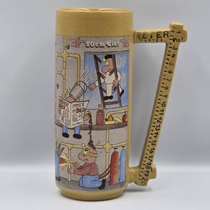 Op de afbeelding: Een gele keramische bierpul met een handvat in de vorm van een liniaal. De pul heeft een cartoonillustratie van een bouwscène met de tekst "20 cm Bier". De handgreep heeft afmetingen in centimeters.