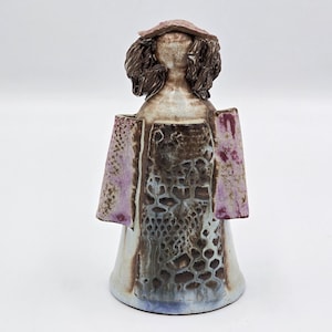 Könnte beinhalten: Keramikfigur einer Person mit Hut und langen Haaren. Die Figur hat ein braunes und violettes Farbschema mit einem strukturierten Muster auf der Vorderseite. Die Figur hat einen ausgestellten Sockel und steht aufrecht.