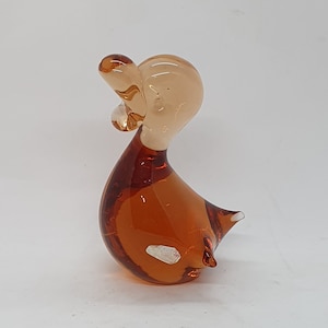 Puede incluir: Una pequeña figurita de pato de vidrio de color ámbar. El pato tiene un cuerpo redondeado y un cuello largo y delgado. La cabeza del pato está inclinada hacia un lado y su pico está abierto.