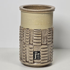 May include: A ceramic vase with a geometric pattern in brown and beige. The vase has a white band around the middle and a black label with the text "JIC VERKSTAD SWEDEN".