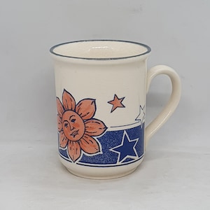 Puede incluir: Una taza de cer&aacute;mica blanca con un dise&ntilde;o azul y naranja que presenta un sol con cara y estrellas.