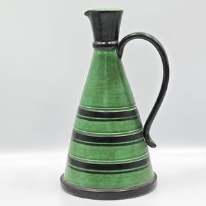 Könnte beinhalten: Eine grüne Keramikkrug mit einem schwarzen Griff und Ausguss. Der Krug hat horizontale schwarze Streifen und einen schwarzen Sockel. Der Griff verläuft von oben zur Seite des Kruges.