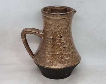 May include: A brown ceramic pitcher with a black base and a textured surface. The pitcher has a handle and a spout.