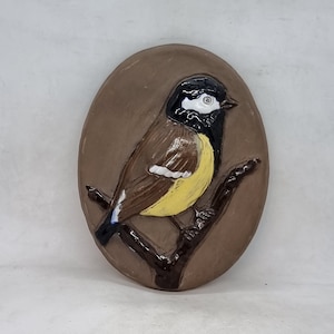 Può includere: Una placca da parete in ceramica con un uccello marrone, giallo e nero appollaiato su un ramo. L'uccello ha una striscia bianca sull'ala e una testa nera con un occhio bianco.