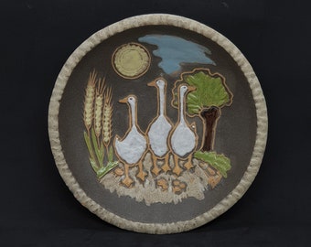 May include: A ceramic plate with a brown background and a white rim. The plate features a painted scene of three white geese walking in a field with green grass, wheat stalks, and a small tree. The sky is blue with a white sun.