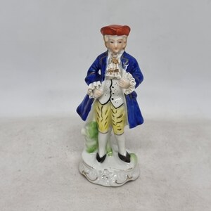 Könnte beinhalten: Eine Porzellanfigur eines Mannes in einem blauen Mantel, einer gelben Hose und einem roten Hut. Er steht auf einem weißen Sockel mit goldenen Akzenten.