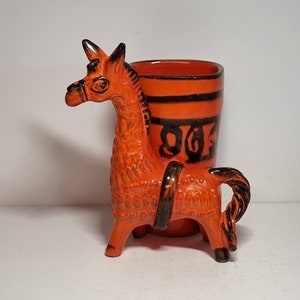 Puede incluir: Taza de cerámica naranja con un diseño en blanco y negro. La taza tiene forma de caballo con una silla de montar negra y una cola negra.