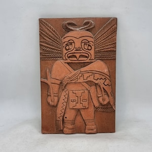 Puede incluir: Un azulejo de terracota con un relieve tallado de una figura estilizada con alas, un tocado de plumas y patrones geométricos. La figura lleva una capa y sostiene un bastón.