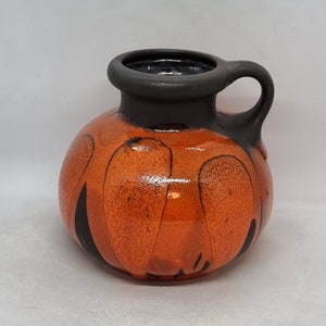 Könnte beinhalten: Eine kleine orangefarbene Keramikvase mit einem schwarzen Henkel und abstrakten schwarzen Mustern. Die Vase hat einen runden Körper und eine leicht ausgestellte Lippe.