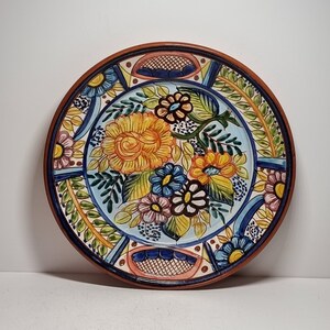 May include: A round ceramic plate with a colorful floral design. The plate features a large yellow rose with smaller pink, blue, and yellow flowers. The plate has a brown rim and a white background.