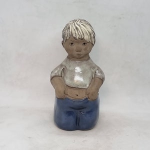 May include: A ceramic figurine of a young child. The figure has light-coloured hair, a grey shirt, and blue trousers. The child's hands are in their pockets. The figurine is on a white background.