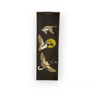 May include: A black rectangular wooden plaque with three painted images of ducks in flight. The ducks are brown, white, and yellow with black accents. The plaque has a yellow circle behind the middle duck.