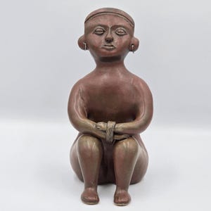 Op de afbeelding: Een bruin kleien beeldje van een zittend figuur met de handen gevouwen voor de knieën. Het figuur heeft een glad, rond hoofd met een hoofdband, opvallende oren met kleine zilverkleurige oorringen en een neutrale gezichtsuitdrukking.