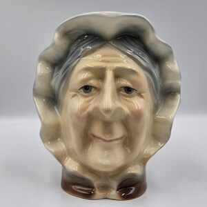 May include: A ceramic figurine of an elderly woman's head, featuring a bonnet and a detailed facial expression. The figurine is painted with realistic skin tones, gray hair, and brown accents. The overall design is a vintage style.