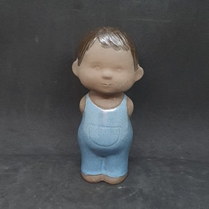 Puede incluir: Una figurita de cerámica de un niño con un mono azul. La figurita tiene la cabeza marrón y el cuerpo marrón claro. La figurita está de pie con las manos juntas delante de ella.