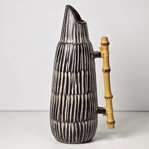Puede incluir: Una jarra alta de cerámica marrón oscuro con rayas blancas verticales y un asa de bambú. La jarra tiene un pico inclinado único y un cuerpo cilíndrico. El asa de bambú está sujeta con cuerda.