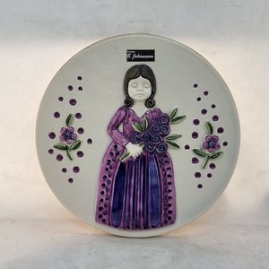 Puede incluir: Un plato de cerámica blanco con un diseño en relieve de una niña con un vestido morado sosteniendo un ramo de flores moradas. El plato tiene un fondo blanco con puntos morados y dos pequeñas flores moradas con tallos verdes. El plato tiene una pequeña etiqueta blanca con el texto "Design B. Johansson".